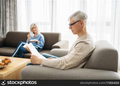 Adult love couple at home in the morning, man reads newspaper, woman drinks coffee. Mature husband and wife sitting on couch, happy family. Adult love couple at home in the morning