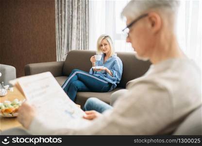 Adult love couple at home in the morning, man reads newspaper, woman drinks coffee. Mature husband and wife sitting on couch, happy family
