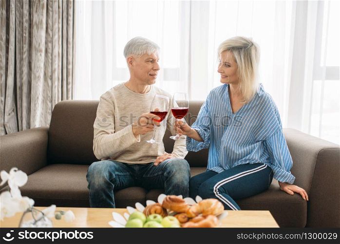 Adult love couple sitting on couch and drinks red wine at home. Mature husband and wife have romantic dinner, happy family