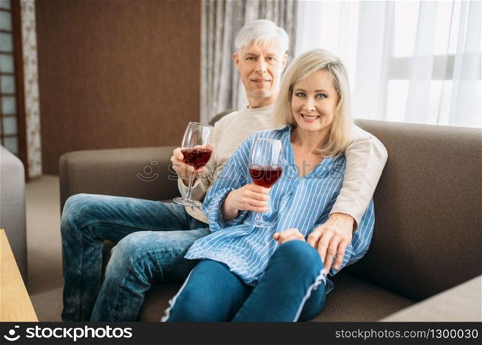 Adult love couple sitting on couch and drinks red wine at home. Mature husband and wife have romantic dinner, happy family. Adult couple sitting on couch and drinks wine