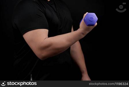 adult male athlete in a black uniform holds purple plastic blue dumbbells in hands, man stands sideways, black background. Bodybuilding and fitness. Healthy active lifestyle