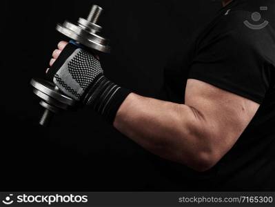 adult male bodybuilder in a black uniform holds in his left hand a steel type-setting dumbbell against a dark background, muscular arm