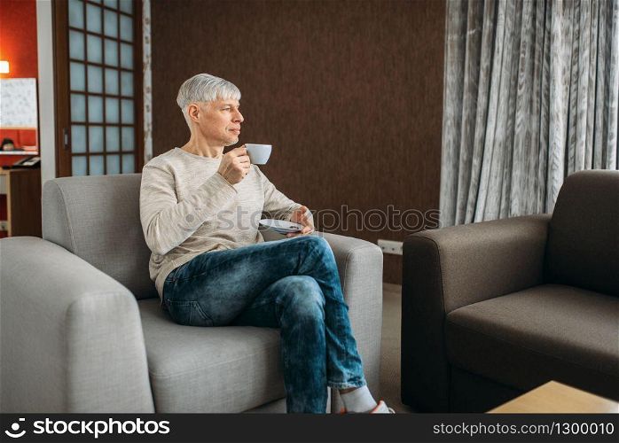 Adult man drinks coffee and reading newspaper at home. Mature male person in jeans relaxes in living room. Adult man drinks coffee and reading newspaper