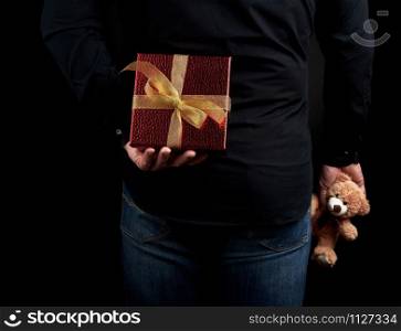 adult man in a black shirt and blue jeans holds a red gift box with a bow behind him, concept of surprise