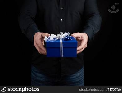 adult man in a black shirt holds a blue square box with a white bow tied on a dark background, concept of congratulations, surprise