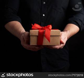 adult man in a black shirt holds in his hand a square brown boxlet tied with a red ribbon on a black background, concept of holiday greetings, surprise and gratitude
