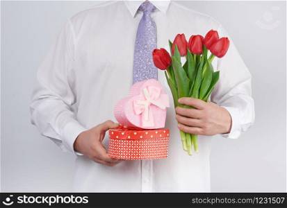 adult man in a white shirt and a lilac tie holding a bouquet of red tulips with green leaves and gift box on a white background, concept for happy birthday, anniversary, valentines day, date