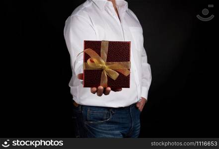 adult man in a white shirt holds a red square box with a knotted golden bow on a dark background, concept of congratulations, surprise
