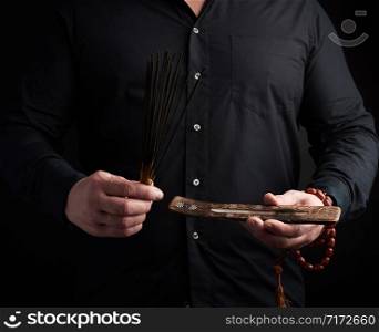 adult man in black clothes holds a stack of incense sticks, dark background