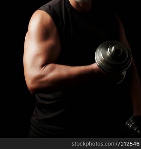 adult man in black clothes holds steel dumbbells in his hands, his muscles are tense, low key, sports background