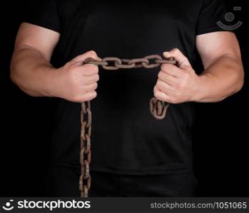 adult man in black clothes stands upright with strained muscles and holds a rusty metal chain, concept of strength and endurance