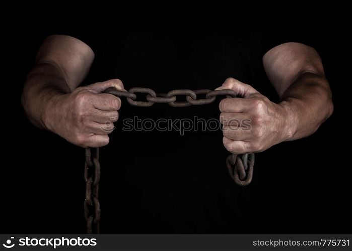 adult man in black clothes stands upright with strained muscles and holds a rusty metal chain, a concept of strength and endurance