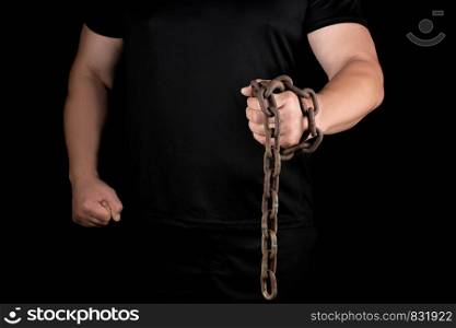 adult man in black clothes stands upright with strained muscles and holds a rusty metal chain, concept of strength and endurance
