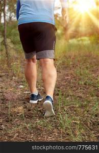 adult man in blue clothes and black shorts runs along the coniferous forest against the bright sun, concept of a healthy lifestyle and running in the fresh air