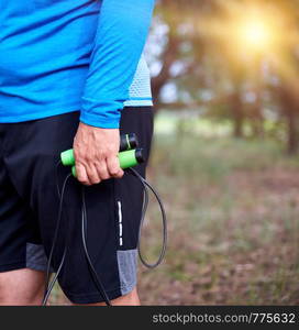 adult man in blue clothes holding a jump rope for playing sports, standing sideways against the setting sun, summer day in the middle of a coniferous forest