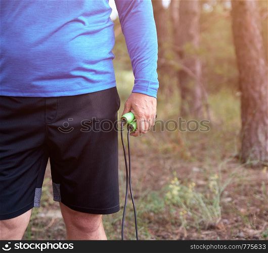 adult man in blue clothes holding a jump rope for playing sports, summer day in the middle of a coniferous forest