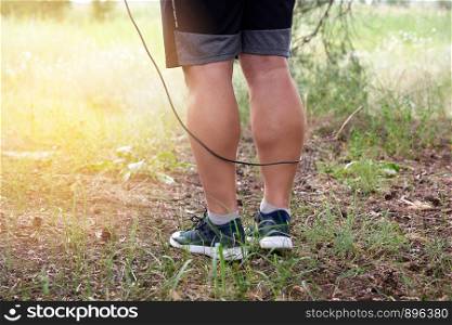 adult man in blue clothes holding a jump rope for playing sports, standing sideways against the setting sun, summer day in the middle of a coniferous forest