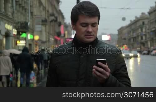 Adult man in the jacket using smartphone in the evening on the urban street on rainy day