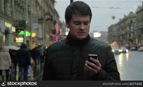 Adult man in the jacket using smartphone in the evening on the urban street on rainy day