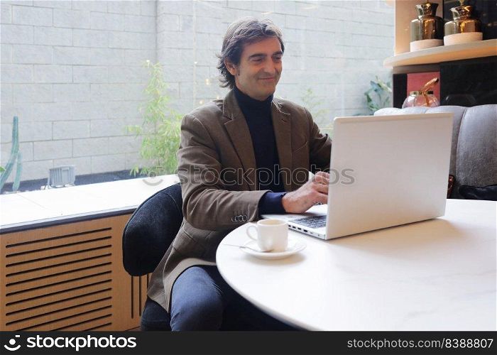 Adult man sitting in a coffee shop while drinking a cup of coffee and using a laptop