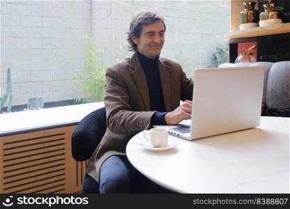 Adult man sitting in a coffee shop while drinking a cup of coffee and using a laptop