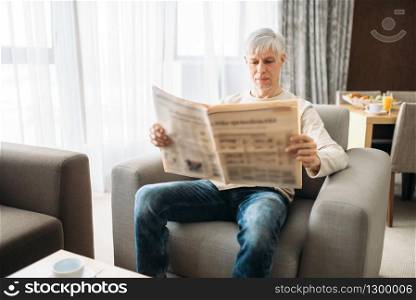 Adult man sitting on couch and reading newspaper at home. Mature male person in jeans relax in armchair