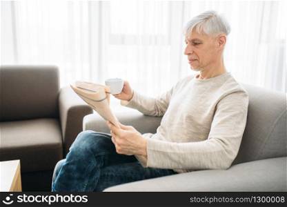 Adult man sitting on couch and reading newspaper at home. Mature male person in jeans relax in armchair. Adult man sitting on couch and reading newspaper