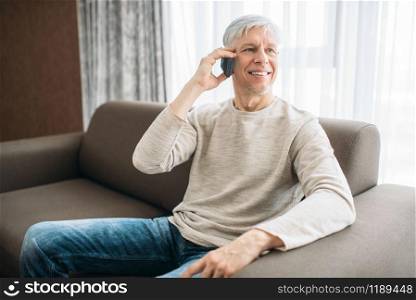 Adult man sitting on couch at home and talking by mobile phone. Mature male person in jeans relax in armchair