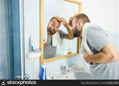 Adult man standing in front of the bathroom mirror brushing his short hair using comb. Guy investigating his receding hairline. Man using comb in bathroom
