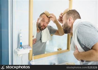 Adult man standing in front of the bathroom mirror brushing his short hair using comb. Guy investigating his receding hairline. Man using comb in bathroom