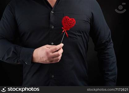 adult man stands on a dark background wearing a black shirt and holding a red carved heart near his chest in his right hand, concept of love