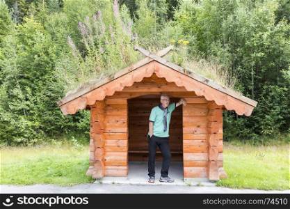 adult man wating for the bus on busstop on the side of the road on the country side in norway. traditional wooden bus stop with grass covered roof,. adult man wating for the bus at traditional bus stop in Norway