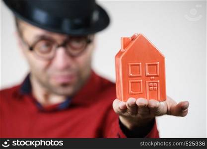 Adult man wearing funny hat and eyeglasses holding small red house model. Guy being real estate agent. Home ownership concept.. Man wearing funny eyeglasses holding house