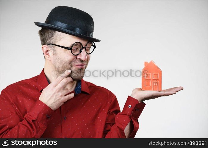 Adult man wearing funny hat and eyeglasses holding small red house model. Guy thinking of becoming real estate agent. Home ownership concept.. Man wearing funny eyeglasses holding house