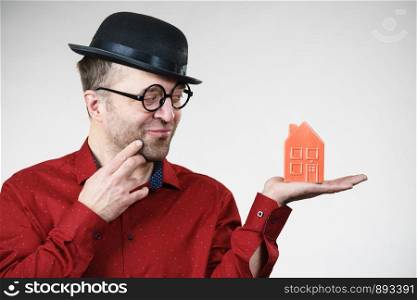 Adult man wearing funny hat and eyeglasses holding small red house model. Guy thinking of becoming real estate agent. Home ownership concept.. Man wearing funny eyeglasses holding house