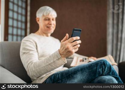 Adult man with phone and newspaper sitting on couch at home. Mature male person in jeans relax in armchair. Adult man with phone sitting on couch at home