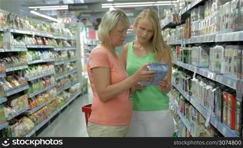 Adult mother and daughter are standing by the supermarket shelf, reading the labels on the packs and talking.