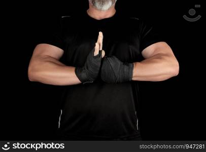 adult muscular athlete in black clothes with rewound hands with a black bandage joined his hands in front of his chest, black background