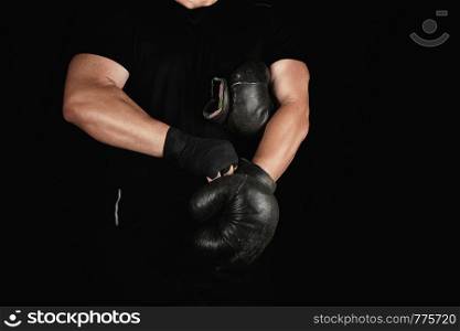 adult muscular man in black clothes puts on leather black boxing gloves on his hands before a competition, his hands are wrapped in a black sports bandage.