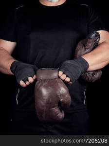 adult muscular man in black clothes puts on leather brown boxing gloves on his hands before a competition, his hands are wrapped in a black sports bandage