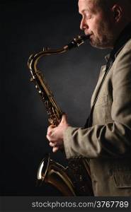 Adult musician playing tenor saxophone, profile view, dark background