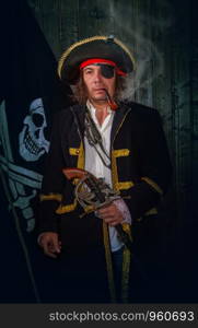 Adult pirate captain in a traditional costume and with a weapon smokes a pipe against the background of a jolly roger. Mature Pirate Capitan