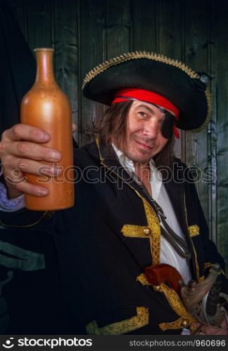 Adult pirate captain in a traditional costume and with weapons drinks rum from a clay bottle against the background of a jolly roger. Mature Pirate Capitan