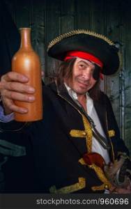 Adult pirate captain in a traditional costume and with weapons drinks rum from a clay bottle against the background of a jolly roger. Mature Pirate Capitan