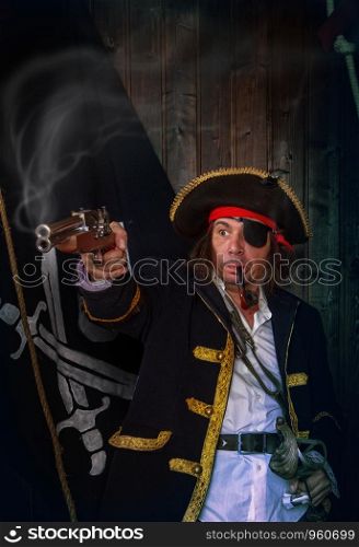 Adult pirate captain in a traditional costume shoots a pistol and holds a sword in his hands against the background of a jolly roger. Mature Pirate Capitan