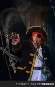 Adult pirate captain in a traditional costume shoots a pistol and holds a sword in his hands against the background of a jolly roger. Mature Pirate Capitan