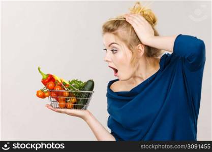 Adult woman do not like to eat raw food, questioning healthy lifestyle recommendations, origin vegetagles. Female holding small shopping basket with products, displeased shocked face expression. Woman with vegetables, shocked face expression