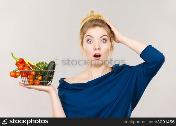 Adult woman do not like to eat raw food, questioning healthy lifestyle recommendations, origin vegetagles. Female holding small shopping basket with products, displeased shocked face expression. Woman with vegetables, shocked face expression