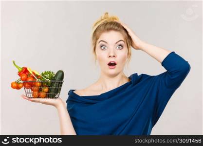 Adult woman do not like to eat raw food, questioning healthy lifestyle recommendations, origin vegetagles. Female holding small shopping basket with products, displeased shocked face expression. Woman with vegetables, shocked face expression