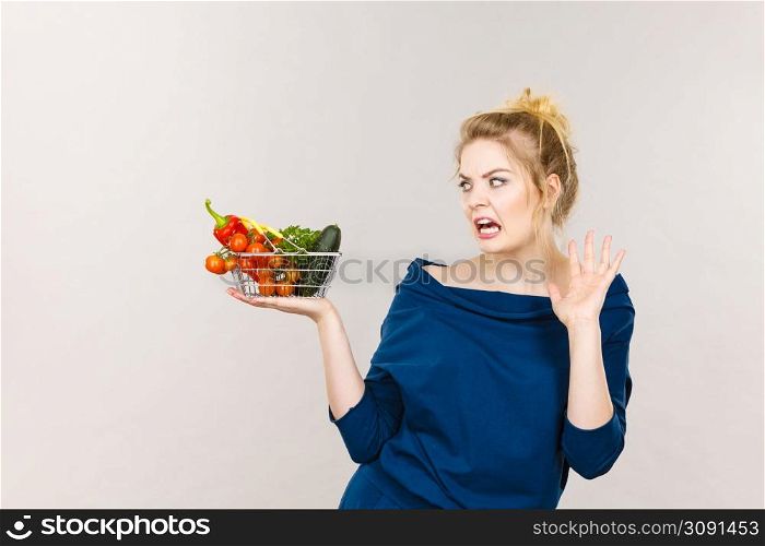Adult woman do not like to eat vegetables, healthy food, vegetarian products. Female holding small shopping basket with green red vegetables, negative displeased face expression, on grey. Woman with vegetables, negative face expression